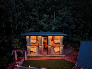 Cabaña de dos pisos con una escalera central, rodeada por un bosque denso.
