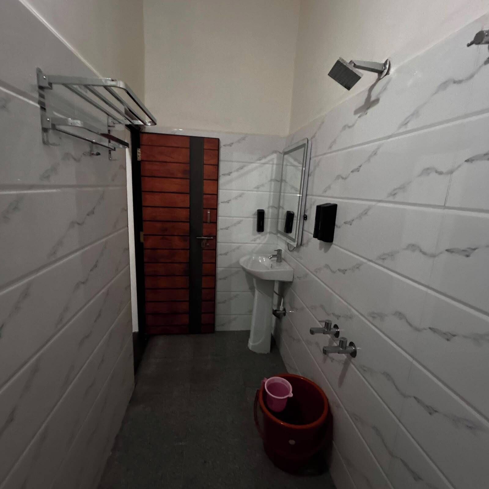 A bathroom with a marble-tiled wall, a wooden door, a sink, a mirror, and a red bucket.