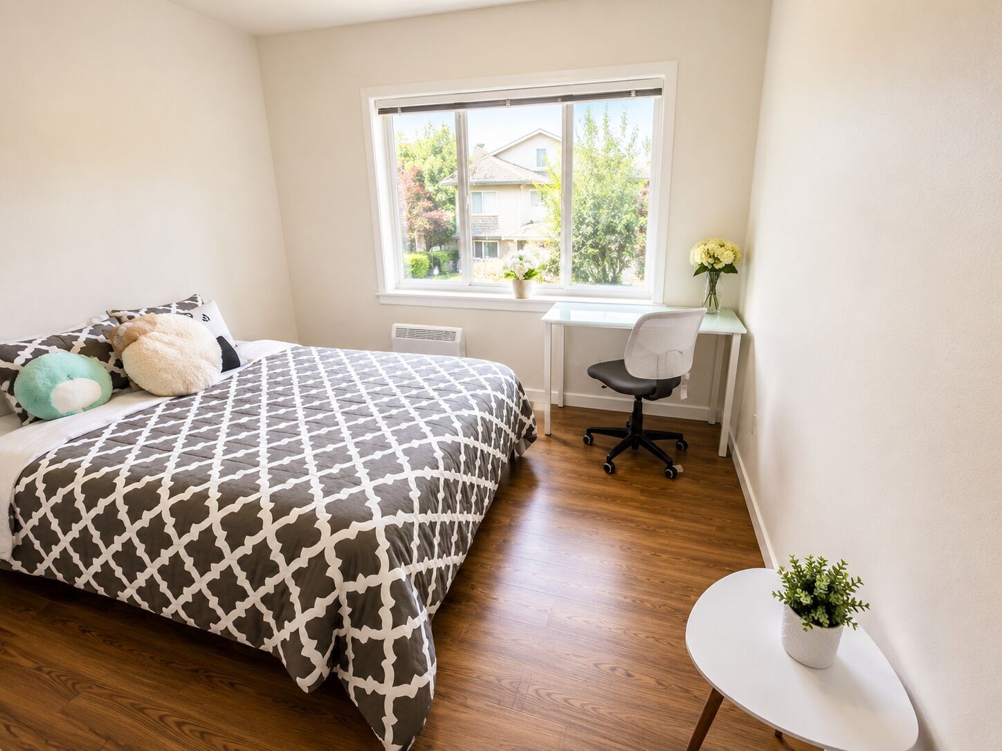A bedroom with a bed, a desk, a chair, a window, and a small table with a plant.