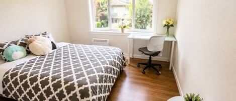 A bedroom with a bed, a desk, a chair, a window, and a small table with a plant.