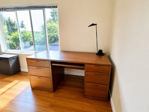 Ein Holztisch mit Schubladen und einer Lampe, nahe einem Fenster mit Blick auf Bäume.