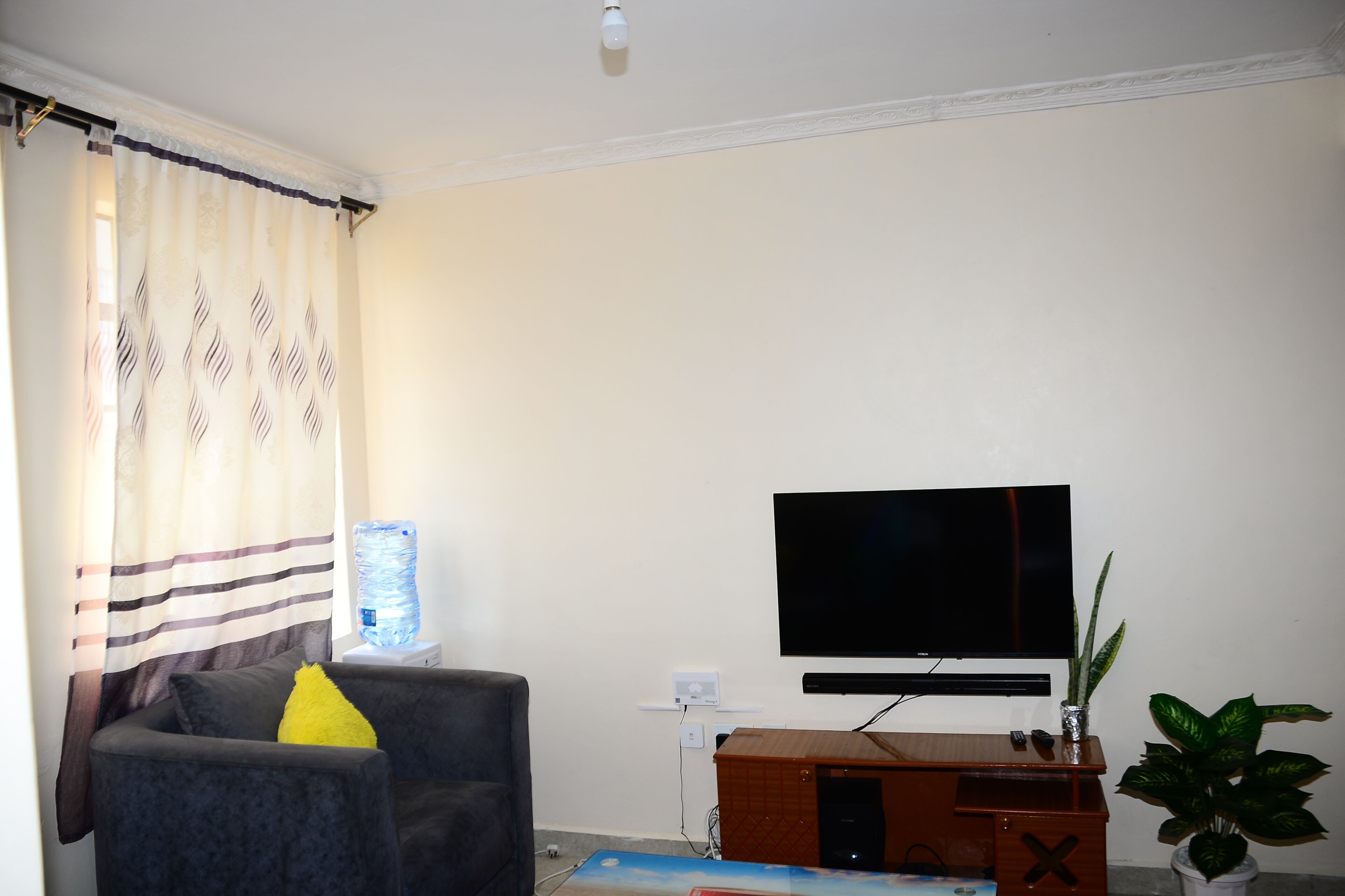 Una sala de estar con un televisor de pantalla plana montado en la pared, un sofá gris con un cojín amarillo, un dispensador de agua y un mueble de madera.