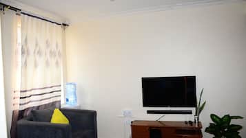 Una sala de estar con un televisor de pantalla plana montado en la pared, un sofá gris con un cojín amarillo, un dispensador de agua y un mueble de madera.