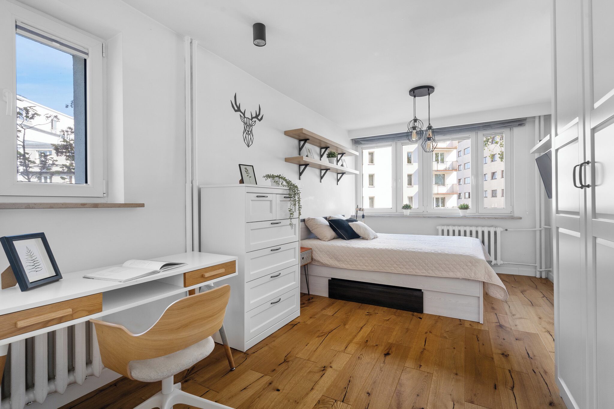 Un dormitorio con cama, escritorio, silla, radiador y ventana con vista a edificios.