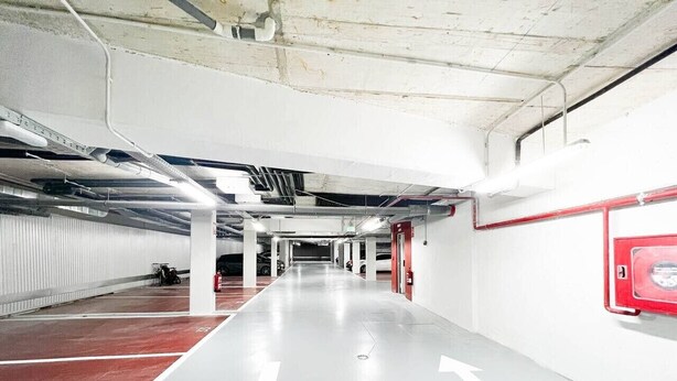 A parking garage with directional arrows on the floor, a fire extinguisher mounted on the wall, and a motorcycle parked in the distance.