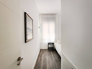 Un couloir étroit mène à une chambre à coucher qui contient un lit, une table de chevet et une fenêtre ornée de rideaux légers.