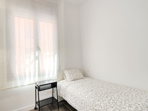 Une petite chambre avec un lit simple, un bureau et une fenêtre ornée de rideaux légers.