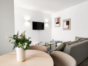 Een moderne woonkamer met een ronde tafel, een bank, een flatscreen televisie aan de muur en twee ingelijste kunstwerken.