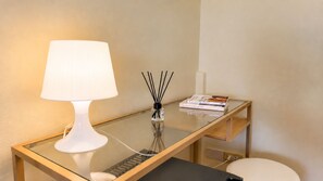 Een tafel met een glazen blad, een witte lamp, een kleine vaas met rietstengels en een stapel boeken.