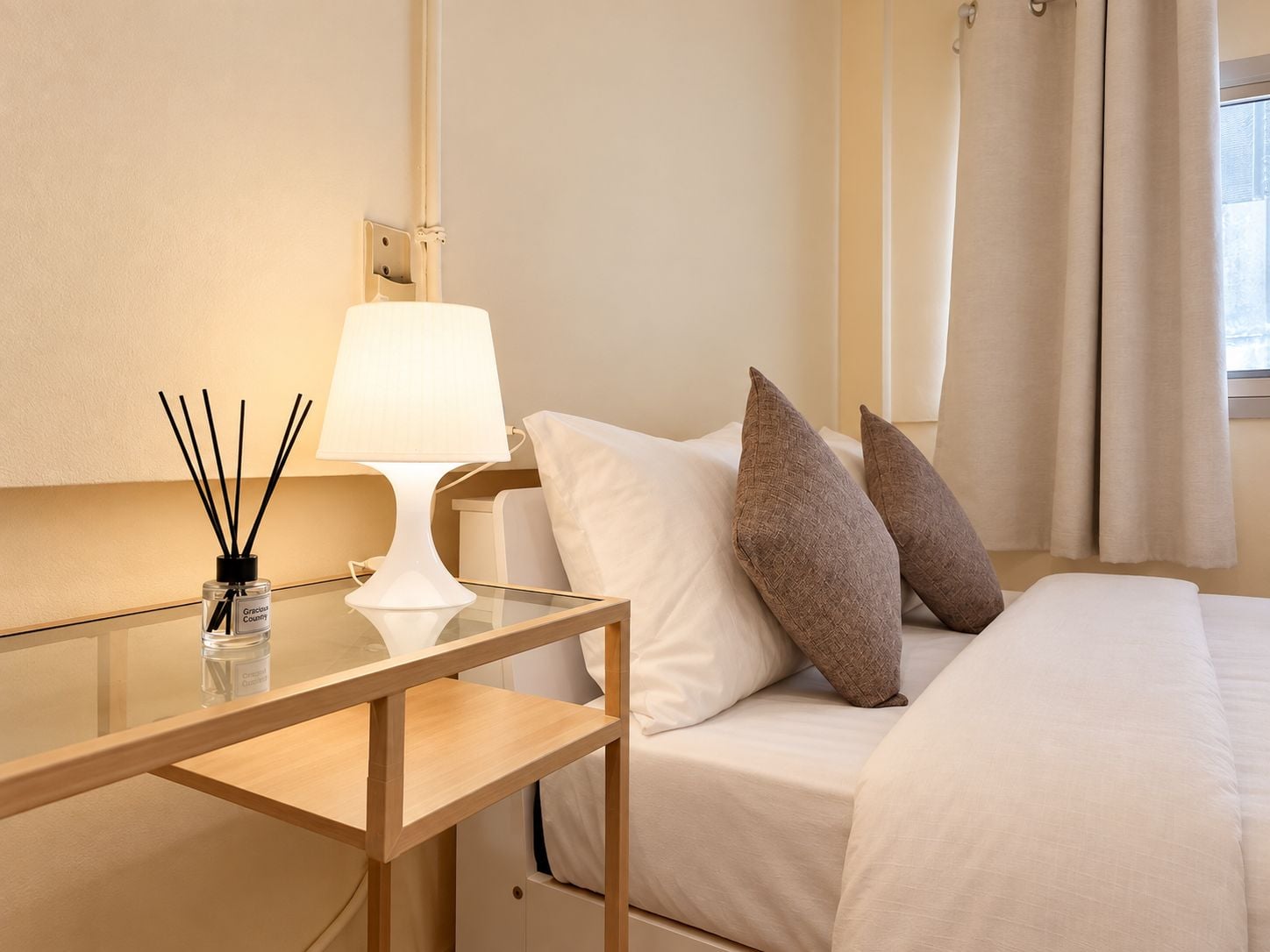 A hotel room with a bed, a bedside table with a lamp, a bottle of reed diffuser, and a window with curtains.