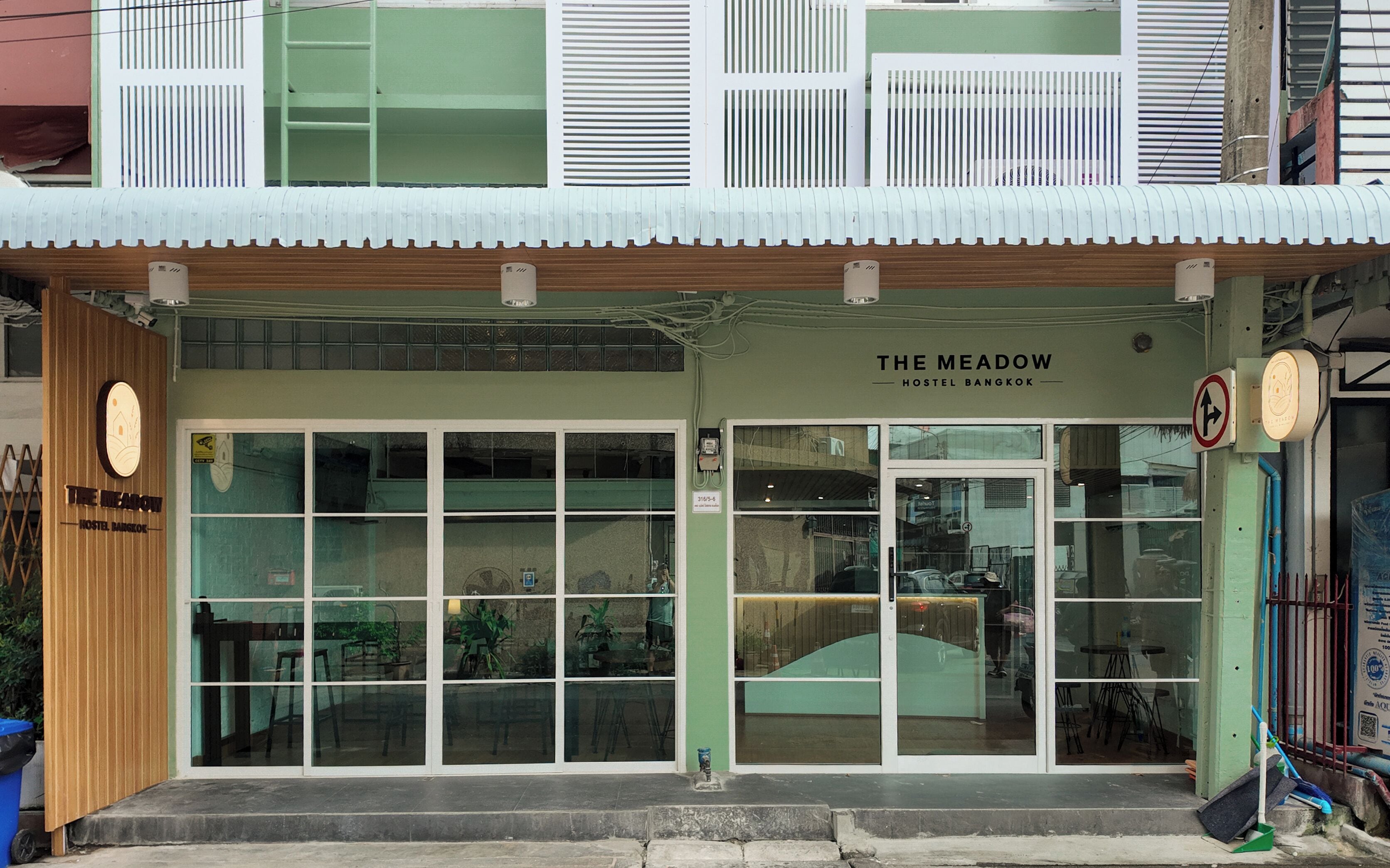 A hostel with a green facade, large windows, and a sign reading 'The Meadow Hostel Bangkok'.
