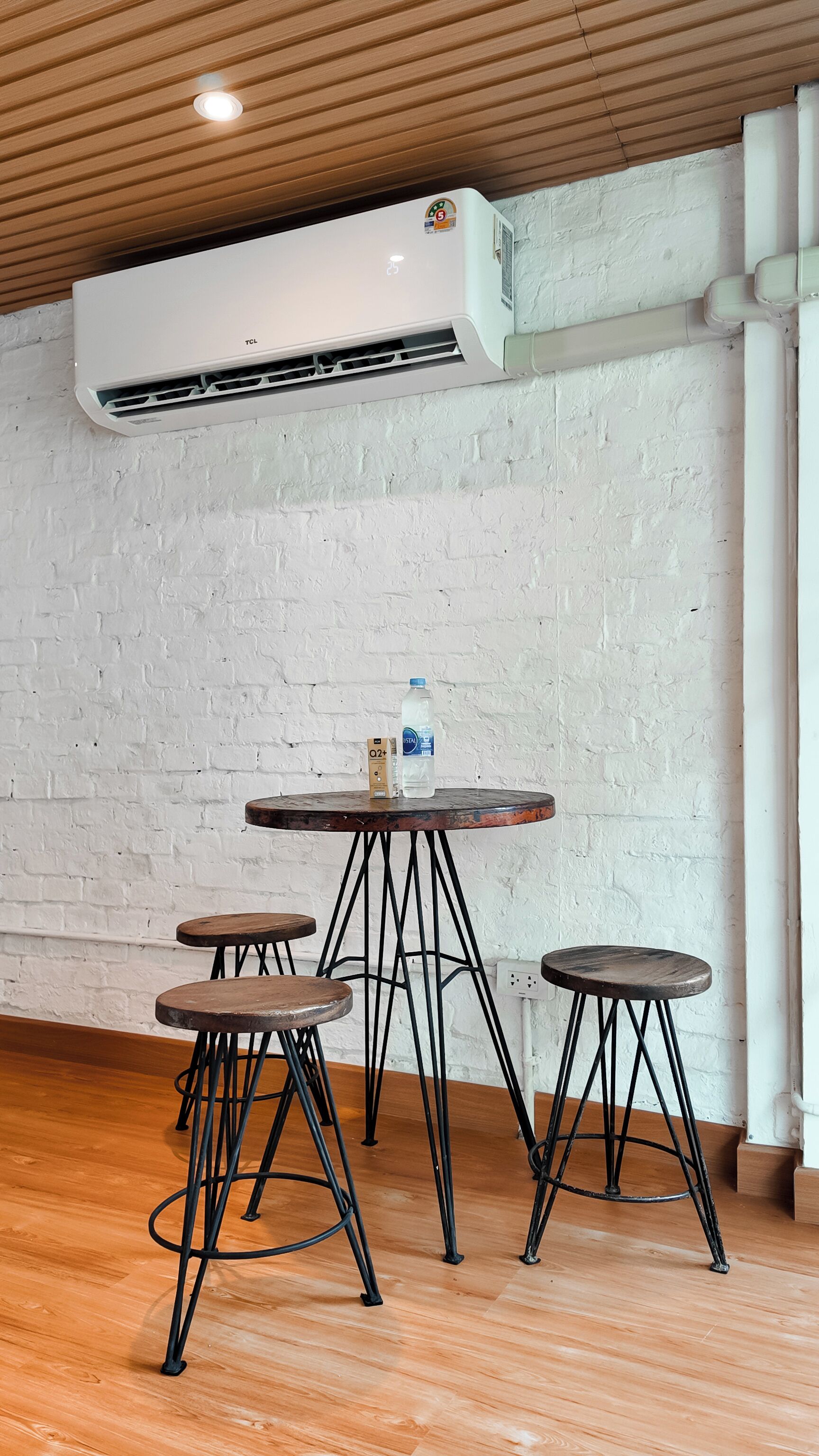 A room with a white brick wall, a round wooden table with four stools, and an air conditioning unit mounted on the ceiling.