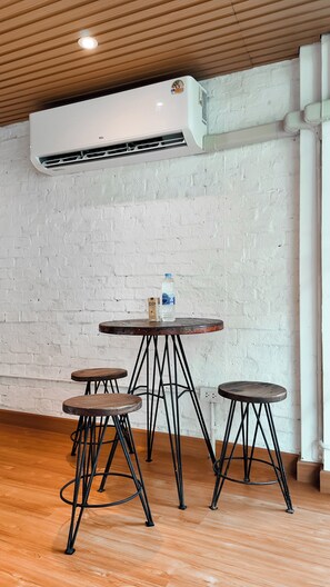 A room with a white brick wall, a round wooden table with four stools, and an air conditioning unit mounted on the ceiling. - The Meadow Hostel Khaosan (Bangkok)