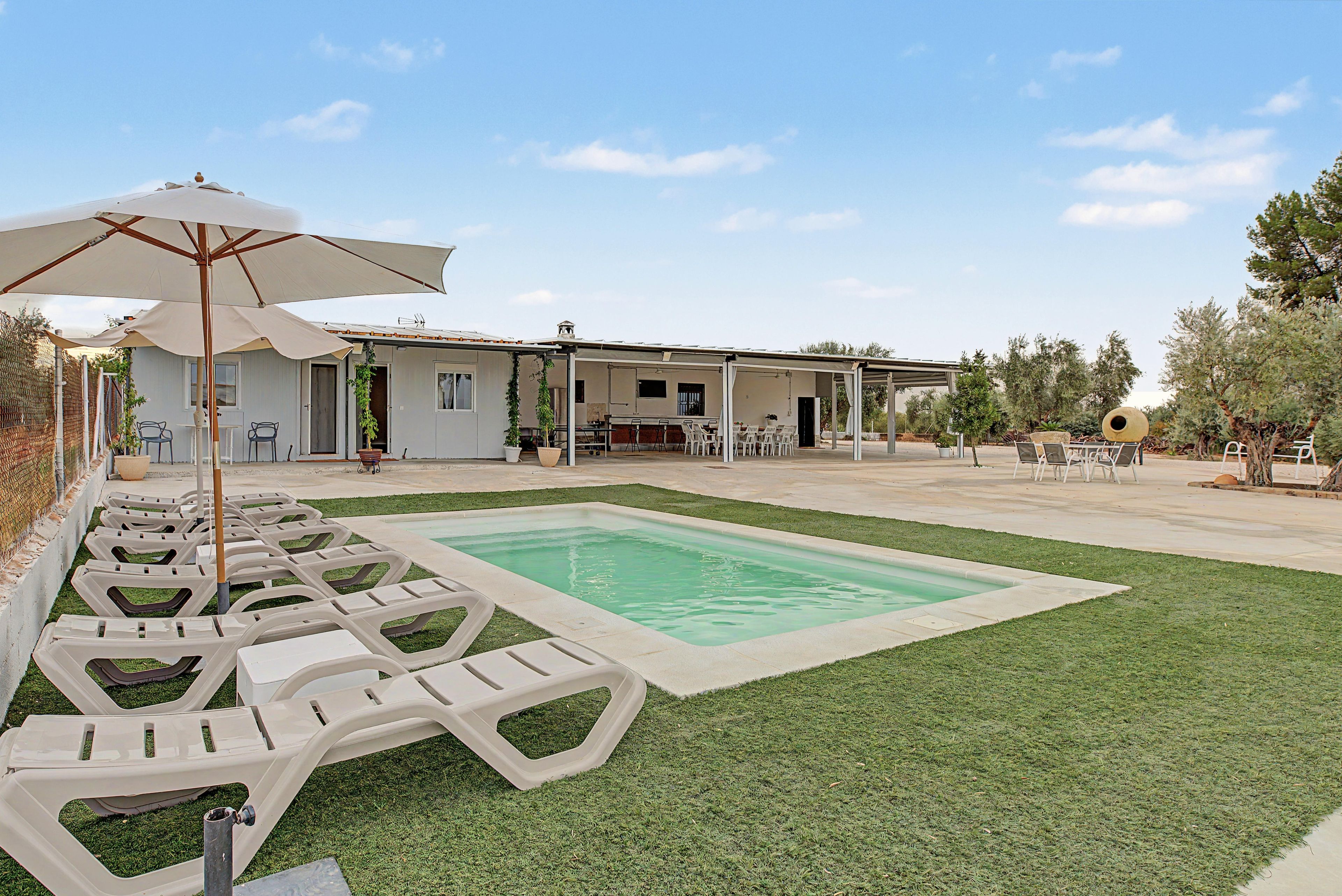 Zona de piscina amb gandules i ombrel·les, un porxo cobert i un edifici amb teulada plana.