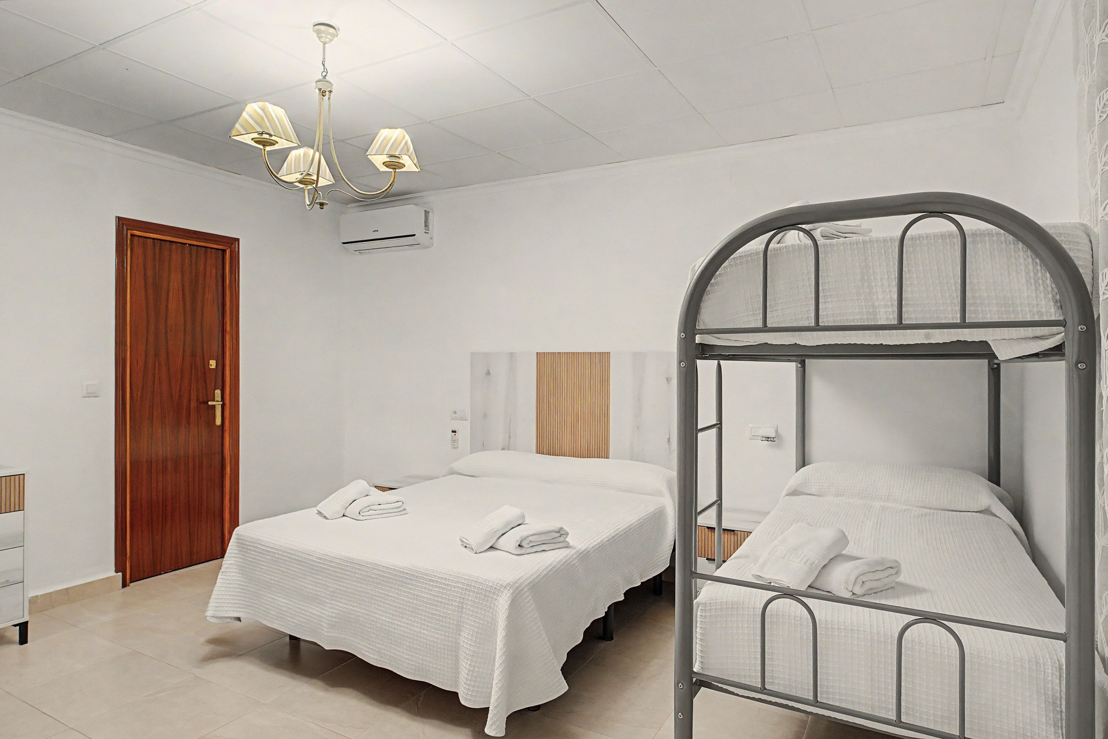 Une chambre avec un lit, une porte en bois, un appareil de climatisation et un lit superposé en métal.