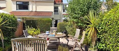 A patio with a round table and chairs, surrounded by greenery and a building in the background.