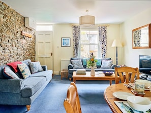 A living room with a stone wall, a sofa with patterned cushions, a coffee table with books, a vase with flowers, and a television on a wooden table. - Finest Retreats | Garston (Salcombe)