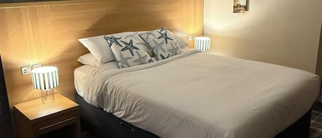 A neatly made bed with a headboard, two bedside lamps, and a small bedside table with a lamp. The room has a checkered floor and a wall with a textured finish.