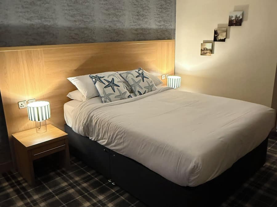 A neatly made bed with a headboard, two bedside lamps, and a small bedside table with a lamp. The room has a checkered floor and a wall with a textured finish.