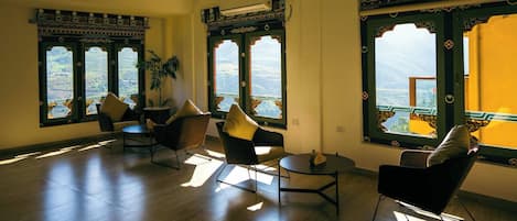 Amplia habitación con un gran ventanal que ofrece vistas a un paisaje montañoso. Hay varias sillas y una mesita, y la habitación presenta un diseño moderno con marcos de ventana estampados.