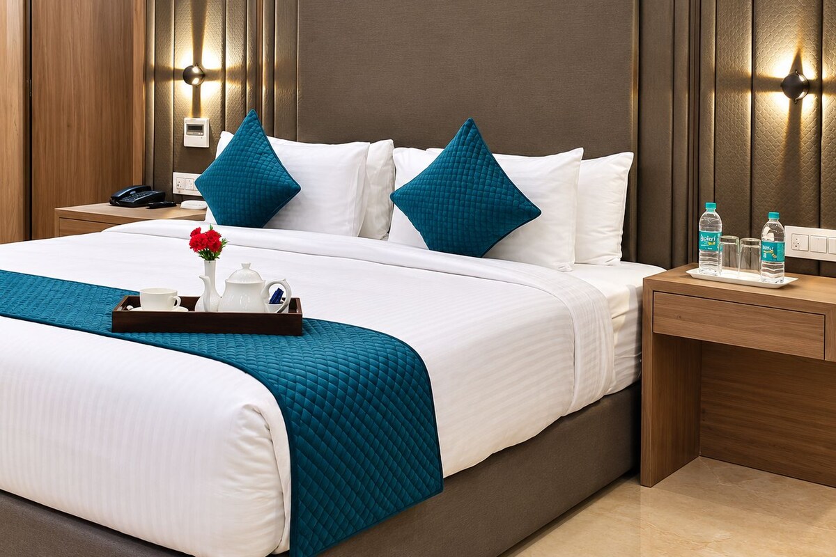 Quarto de hotel com uma cama grande, duas mesas de cabeceira, um telefone, um tabuleiro com chaleira e chávenas, e garrafas de água.