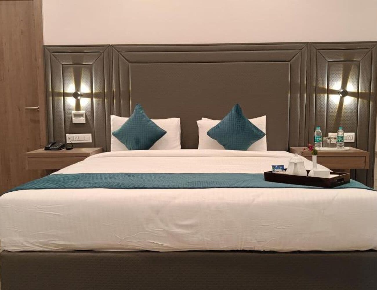 Ein Hotelzimmer mit einem großen Bett, zwei Nachttischen mit Lampen, einem Tablett mit Annehmlichkeiten und einer Holztür.