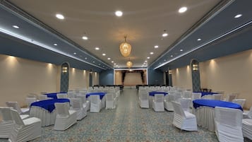 Uma sala de conferências com fileiras de cadeiras e mesas, um tapete estampado e um lustre.