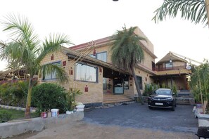 A two-story building with a brick facade, a car parked in front, and palm trees around. - Surya Resort, Peaceful Stay with Modern Comforts (Chittorgarh)