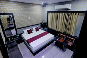 A hotel room with a brick-patterned wall, a large bed with white and red bedding, a bedside table with a lamp, a small table with a chair, and a window with curtains. - Surya Resort, Peaceful Stay with Modern Comforts (Chittorgarh)