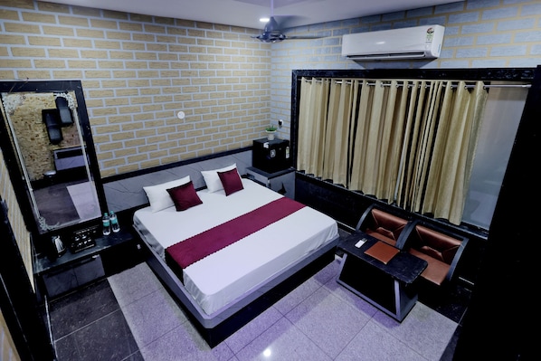 A hotel room with a brick-patterned wall, a large bed with white and red bedding, a bedside table with a lamp, a small table with a chair, and a window with curtains.