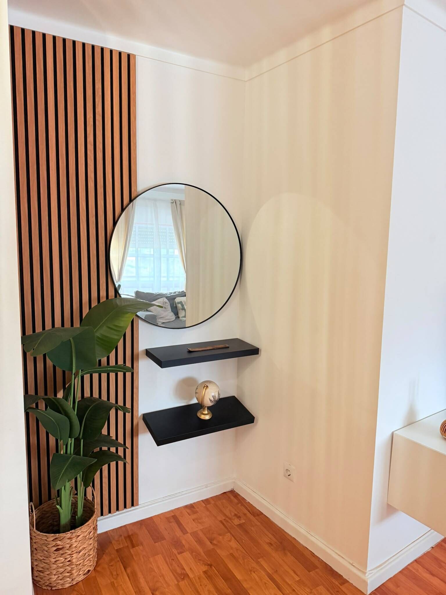 Eine Ecke eines Raumes mit einer Wand aus Holzleisten, einem runden Spiegel, einem kleinen Regal mit einem Globus und einer Topfpflanze.