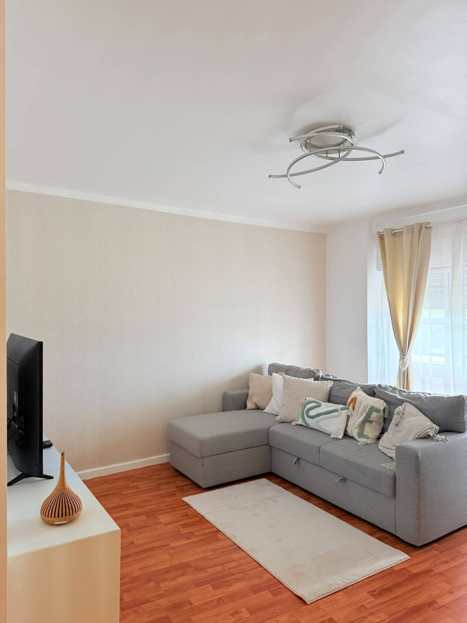 Una sala de estar con un sofá seccional gris, un televisor de pantalla plana, piso de madera y una lámpara en el techo.