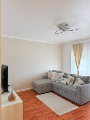 A living room with a grey sectional sofa, a flat-screen TV, a wooden floor, and a ceiling light fixture. - Apart Lisbon Airport III (Lisboa)