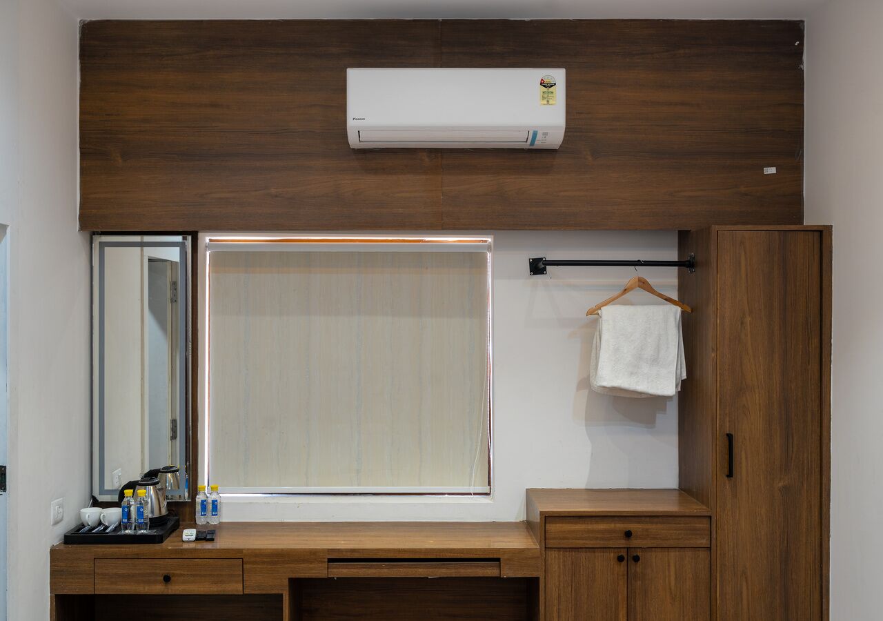 A hotel room with a wooden vanity, a mirror, a window with blinds, a towel hanging on a rod, and an air conditioning unit mounted on the wall.