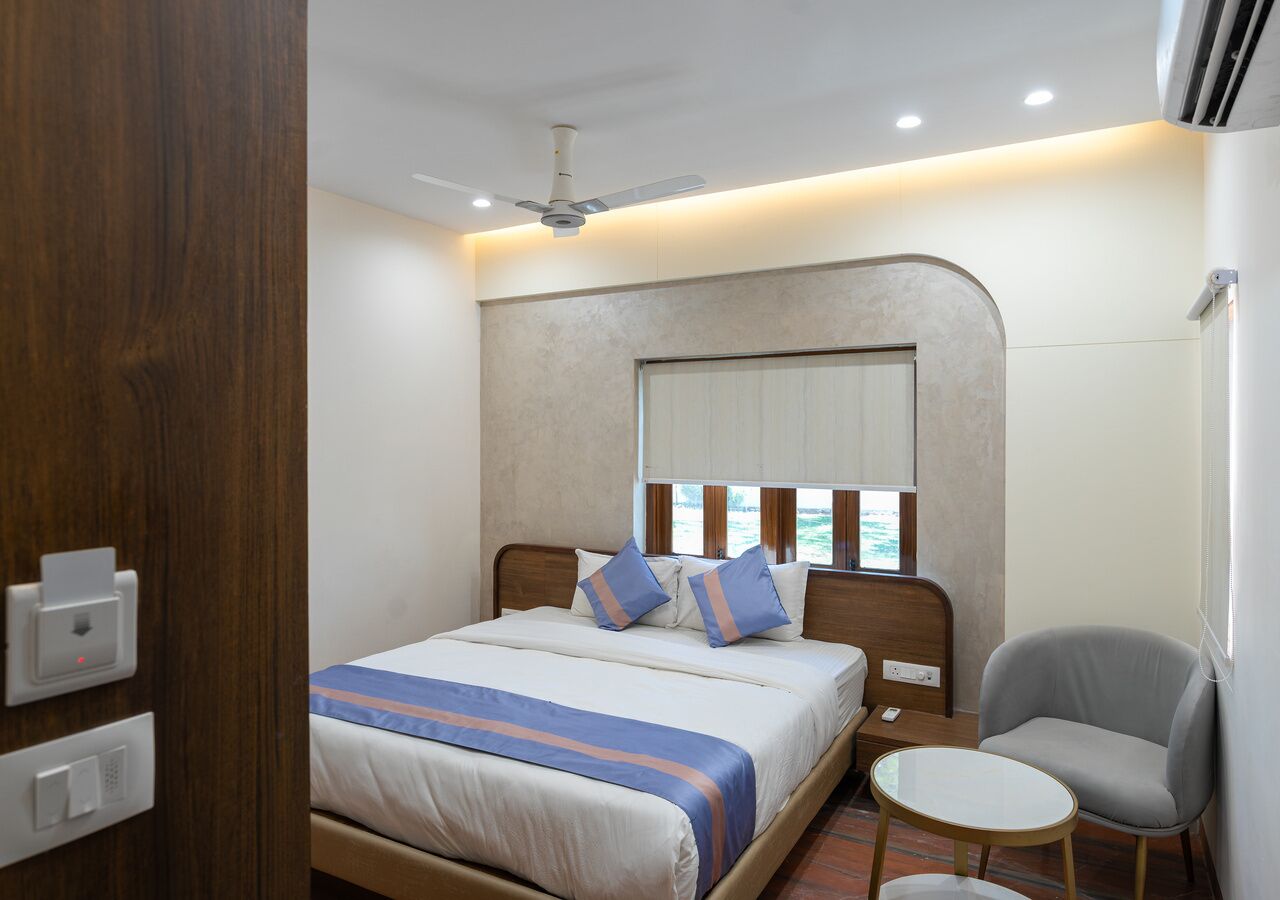 A hotel room with a bed, bedside table, chair, and a window with blinds.