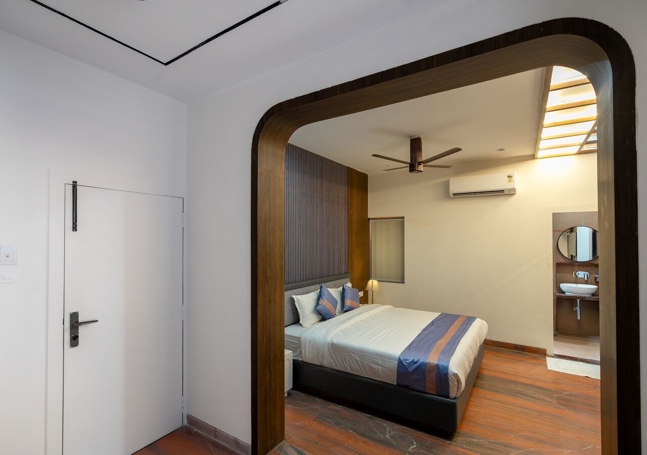 Habitación de hotel con cama, ventilador de techo, equipo de aire acondicionado, baño con lavamanos y espejo, y piso de madera.