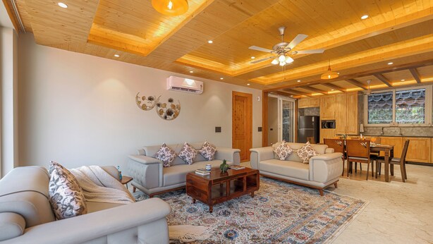 A spacious living room with a high ceiling, wooden beams, a ceiling fan, and a large window with a view of the outdoors.