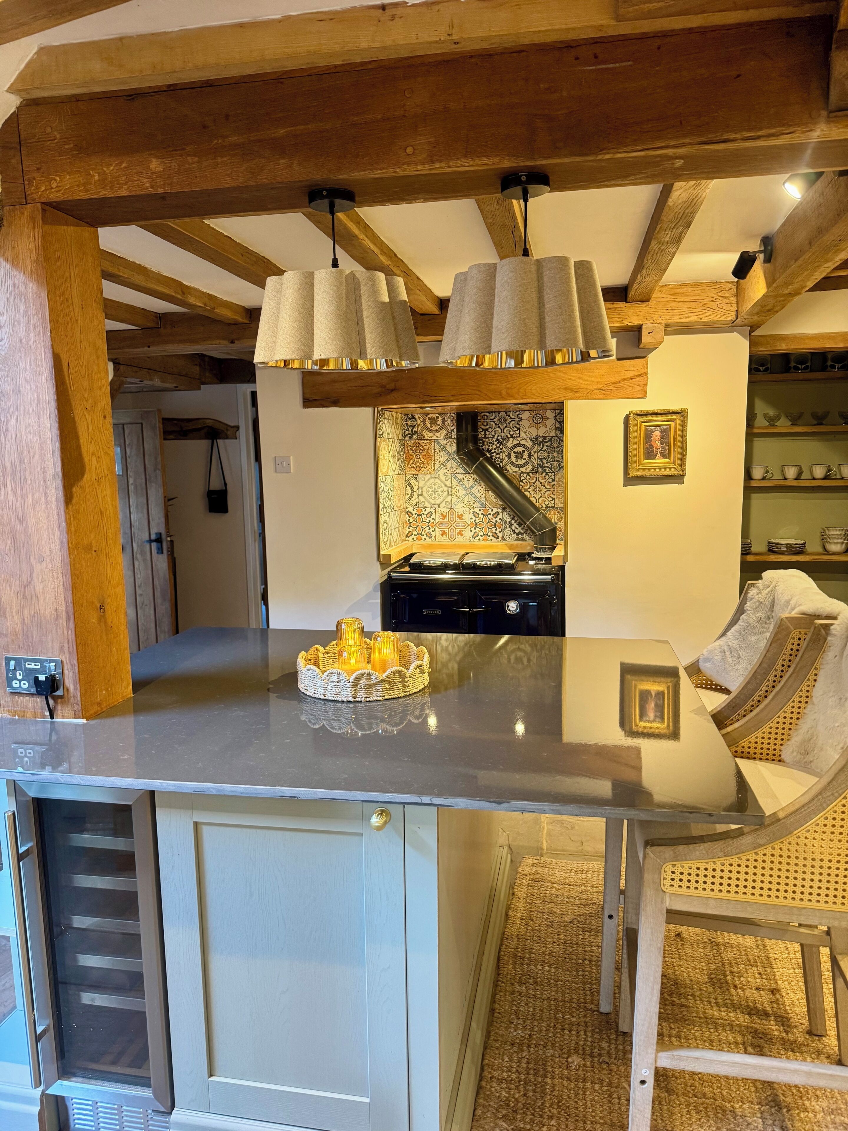 Uma cozinha com ilha central, vigas de madeira, luminárias pendentes e painel de azulejos.