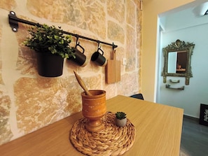A rustic kitchen with a stone wall, a wooden table, a woven placemat, a mortar and pestle, hanging pots, and a mirror. - Patrelaki Stone Residence Chania, with private yard (Chania)