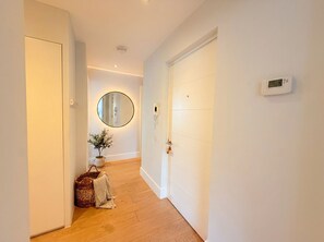 A hallway with a wooden floor, a white door, a round mirror, and a thermostat on the wall. - Modern 2‑Bed Apartment with Balcony, Super‑King Bed & 50m to Swanage Beach (Swanage)