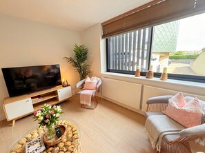 A living room with a flat-screen TV, a sofa, a chair, a coffee table with snacks, and a window with blinds. - Modern 2‑Bed Apartment with Balcony, Super‑King Bed & 50m to Swanage Beach (Swanage)