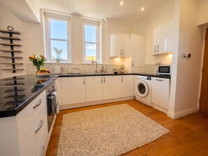 A modern kitchen with white cabinetry, a black countertop, a washing machine, and a window with a view. - Stylish Swanage Apartment | Near Beach & Town (Swanage)