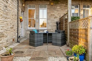 A stone-walled patio with a wicker table and chairs, potted plants, and a wooden fence. - Stylish Swanage Apartment | Near Beach & Town (Swanage)