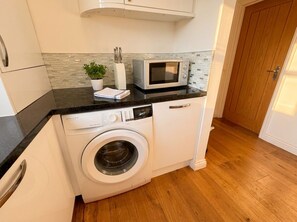 A compact kitchen with a washing machine, a microwave, and a potted plant on the counter. - Stylish Swanage Apartment | Near Beach & Town (Swanage)