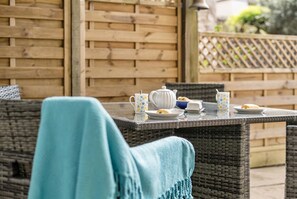 Ein Korbstuhl-Garnitur mit Teekanne, Tassen und einem Teller mit Essen auf einer Terrassenfläche.