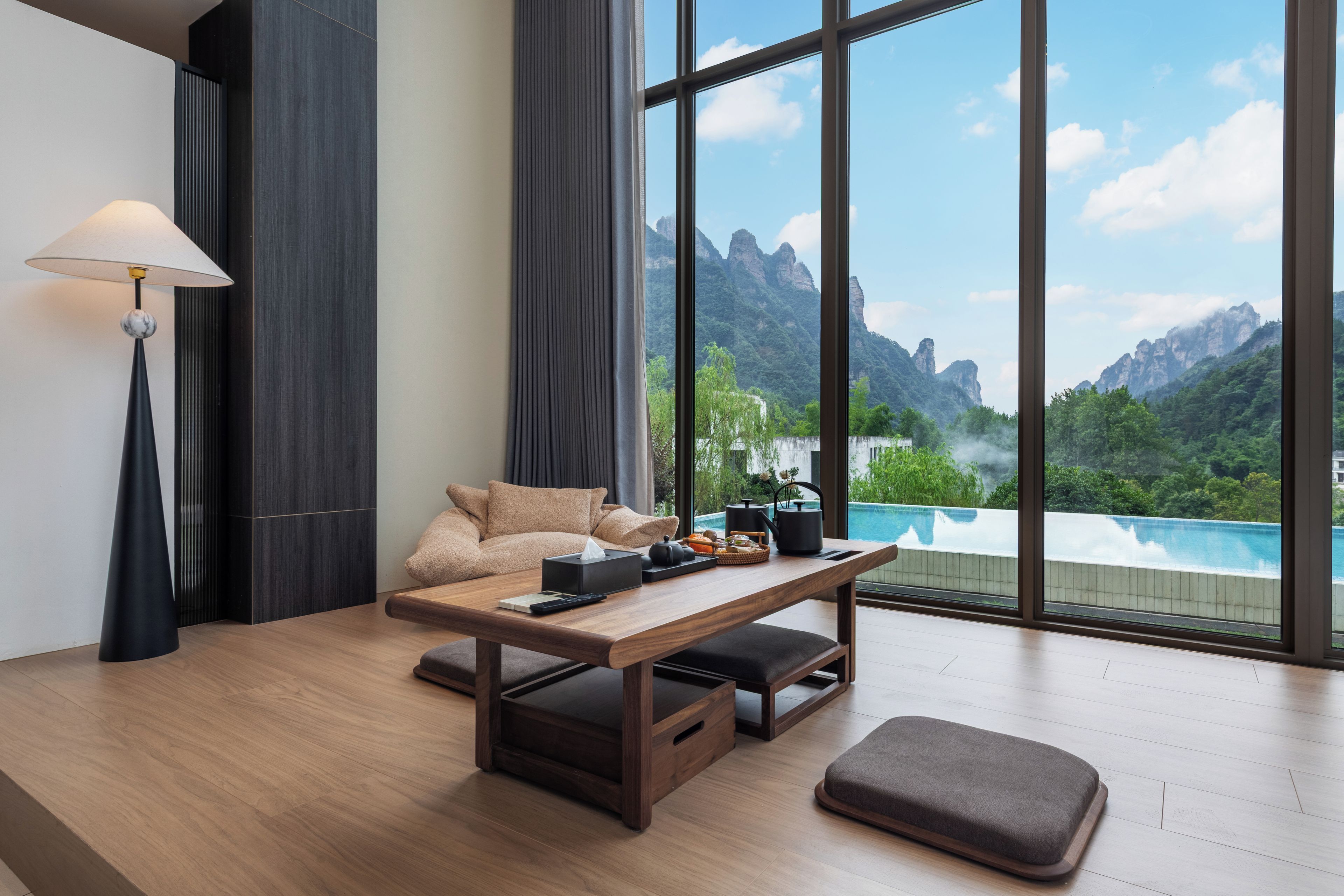 A modern room with a wooden table, a sofa, a lamp, and a view of a mountain landscape through large windows.