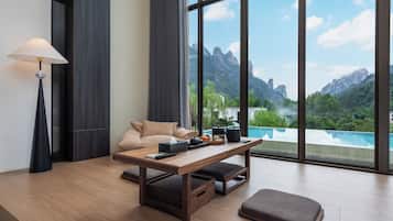 A modern room with a wooden table, a sofa, a lamp, and a view of a mountain landscape through large windows.