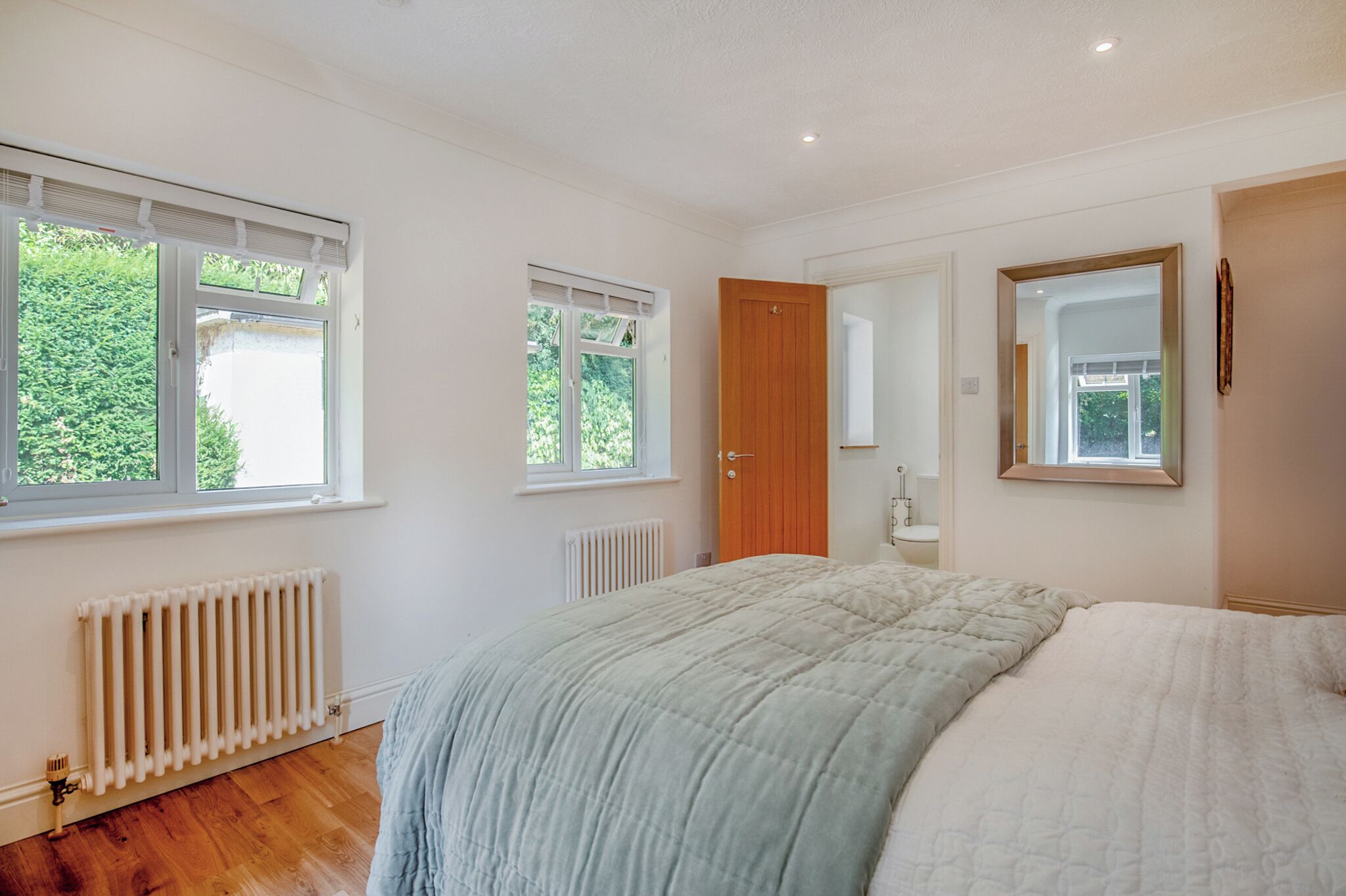 Een slaapkamer met een groot bed, twee ramen, een radiator, een deur en een spiegel.