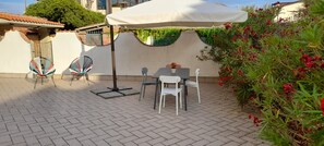 Eine Terrasse im Freien mit Tisch und Stühlen, einem überdachten Bereich und einem Garten mit roten Blumen.
