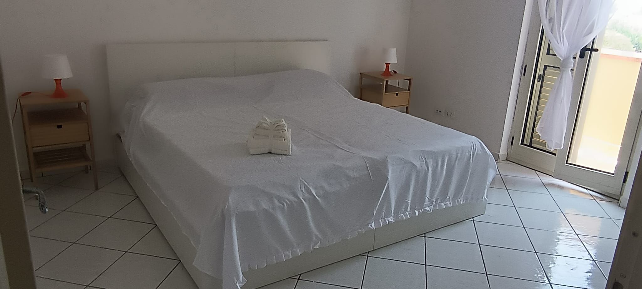 Habitación individual con una cama sencilla cubierta con un edredón blanco, una mesita de noche con lámpara y una ventana con cortinas.